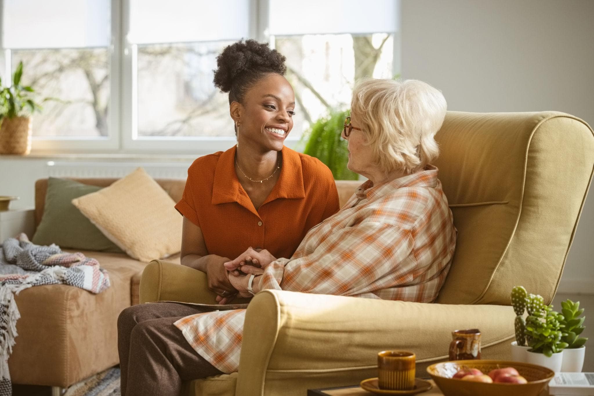 A caregiver holding hands and smiling with an older woman in a warm living room