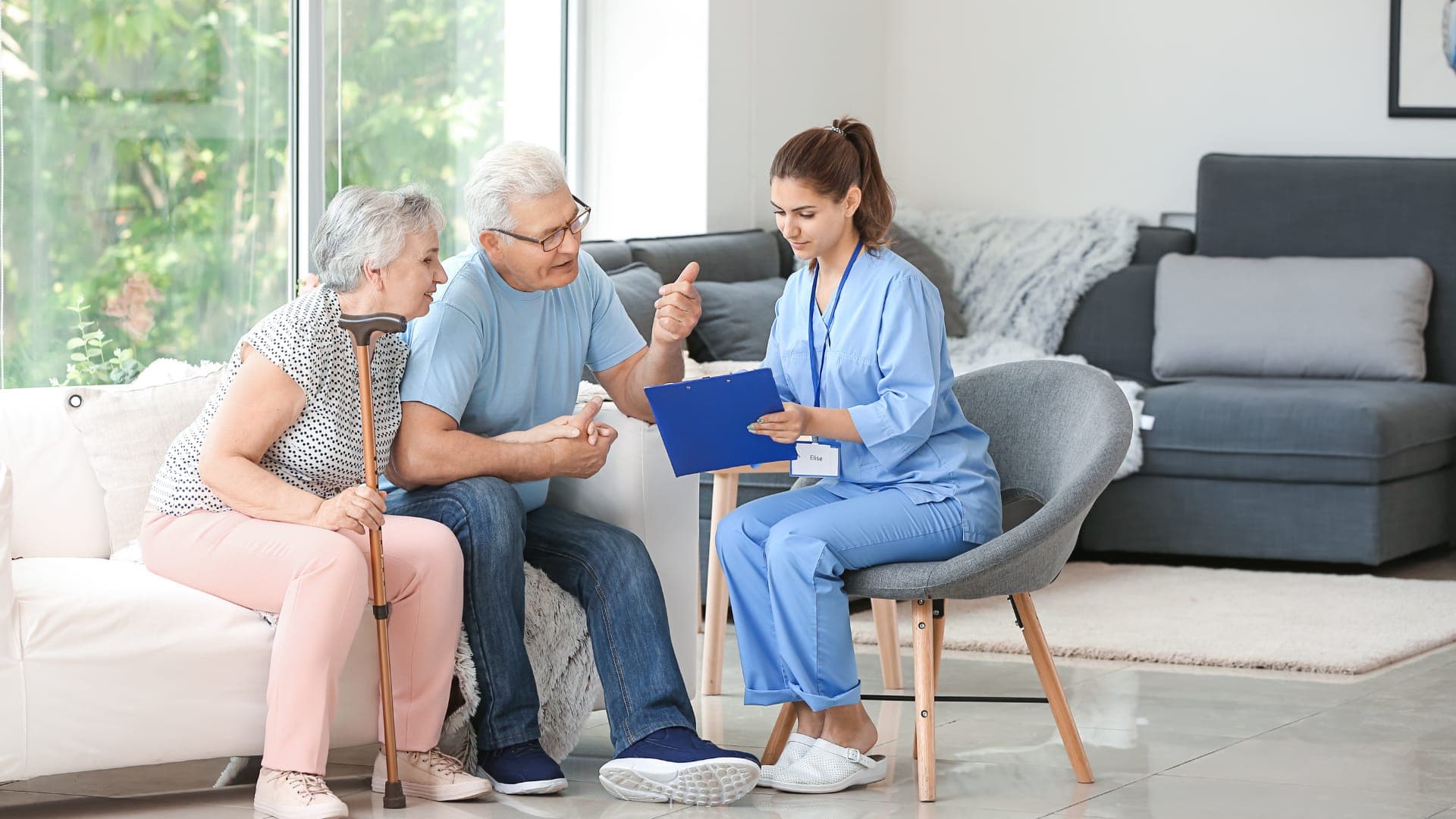 An older couple on their sofa talking with a care coordinator, who sits across from them with a clipboard.